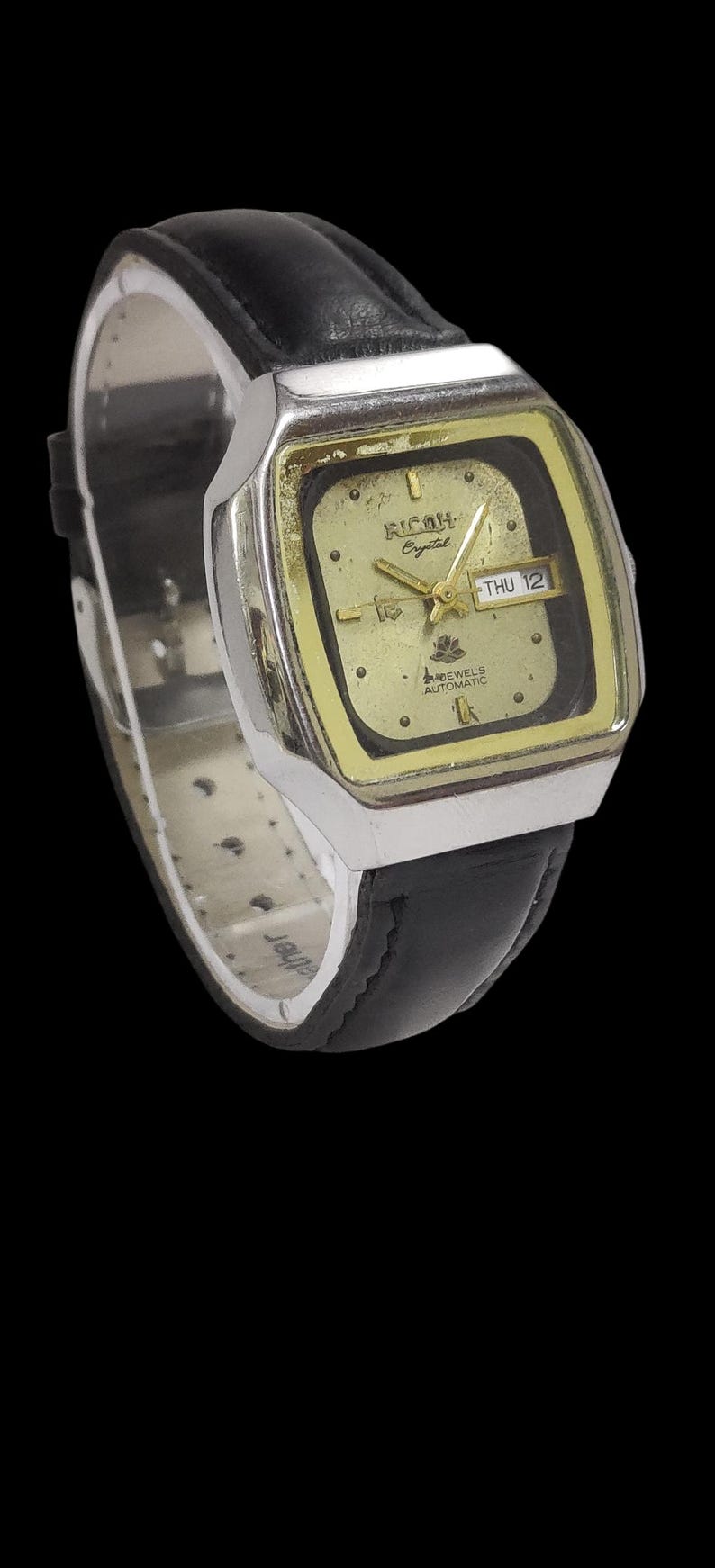 May include: A vintage Ricoh Crystal automatic wristwatch with a gold-toned square face and black leather strap. The watch face displays the day and date, with the text "THU 12". The watch has a silver-toned case.