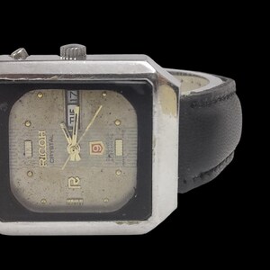 Może przedstawiać: Zegarek Ricoh Crystal w stylu vintage z kwadratową, srebrną kopertą i czarnym sk&oacute;rzanym paskiem. Tarcza zegarka jest w kolorze jasnobeżowym ze złotymi wskaz&oacute;wkami i znacznikami. Wyświetlany jest dzień i data. Zegarek na czarnym tle.