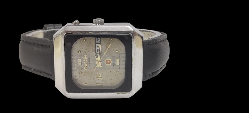 Może przedstawiać: Zegarek Ricoh Crystal w stylu vintage z kwadratową, srebrną kopertą i czarnym sk&oacute;rzanym paskiem. Tarcza zegarka jest w kolorze jasnobeżowym ze złotymi wskaz&oacute;wkami i znacznikami. Wyświetlany jest dzień i data. Zegarek na czarnym tle.
