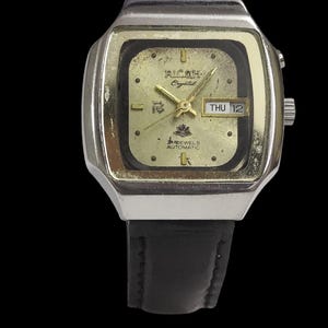 May include: A vintage Ricoh Crystal automatic watch with a gold-tone square face and black leather strap. The watch face shows the day and date, with the text "Ricoh Crystal" and "12 Jewels Automatic".