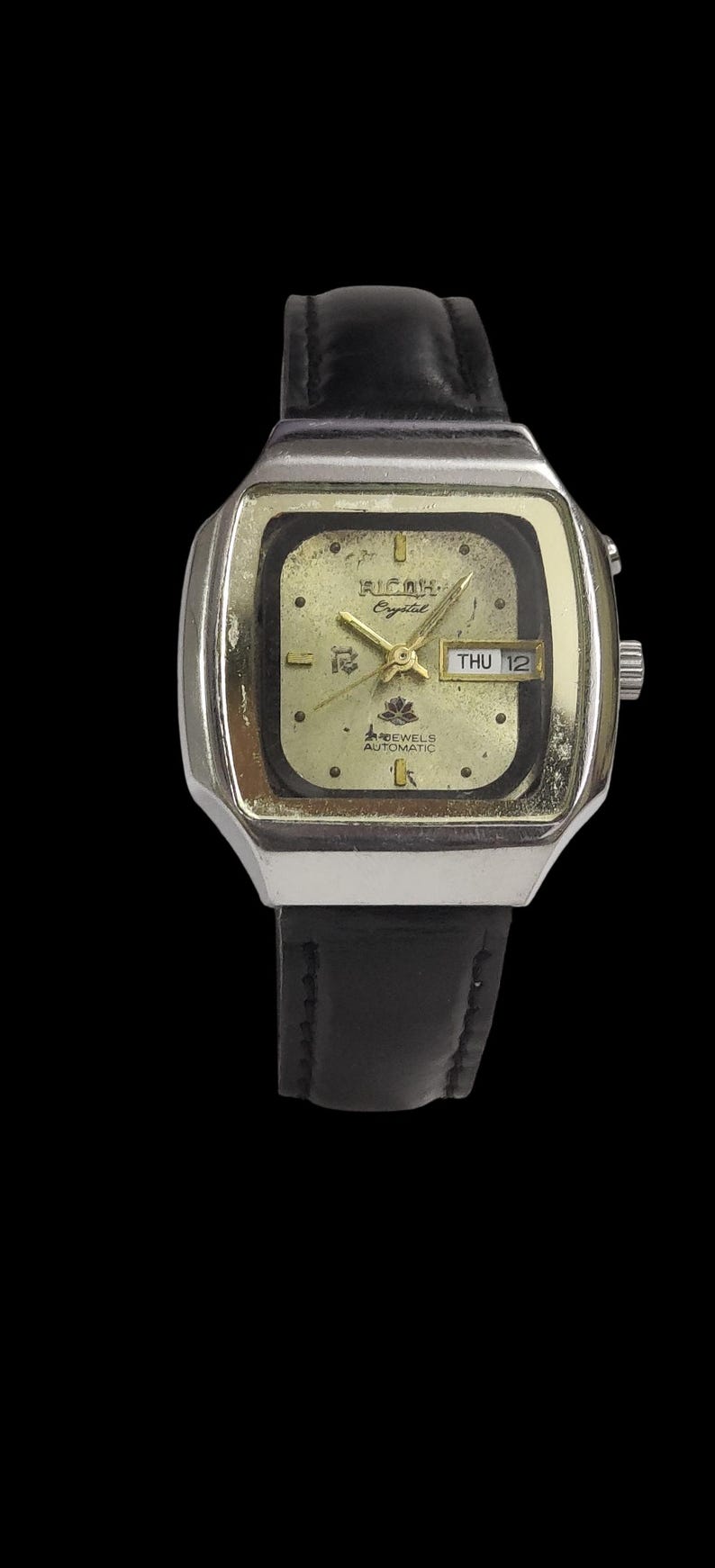 May include: A vintage Ricoh Crystal automatic watch with a gold-tone square face and black leather strap. The watch face shows the day and date, with the text "Ricoh Crystal" and "12 Jewels Automatic".