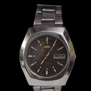 K&ouml;nnte beinhalten: Silberfarbene Orient-Uhr mit dunkelbraunem Zifferblatt. Die Uhr verf&uuml;gt &uuml;ber eine Tages- und Datumsanzeige, Leuchtmarkierungen und ein silbernes Metallarmband. Auf dem Zifferblatt steht "Orient" und "Crystal".