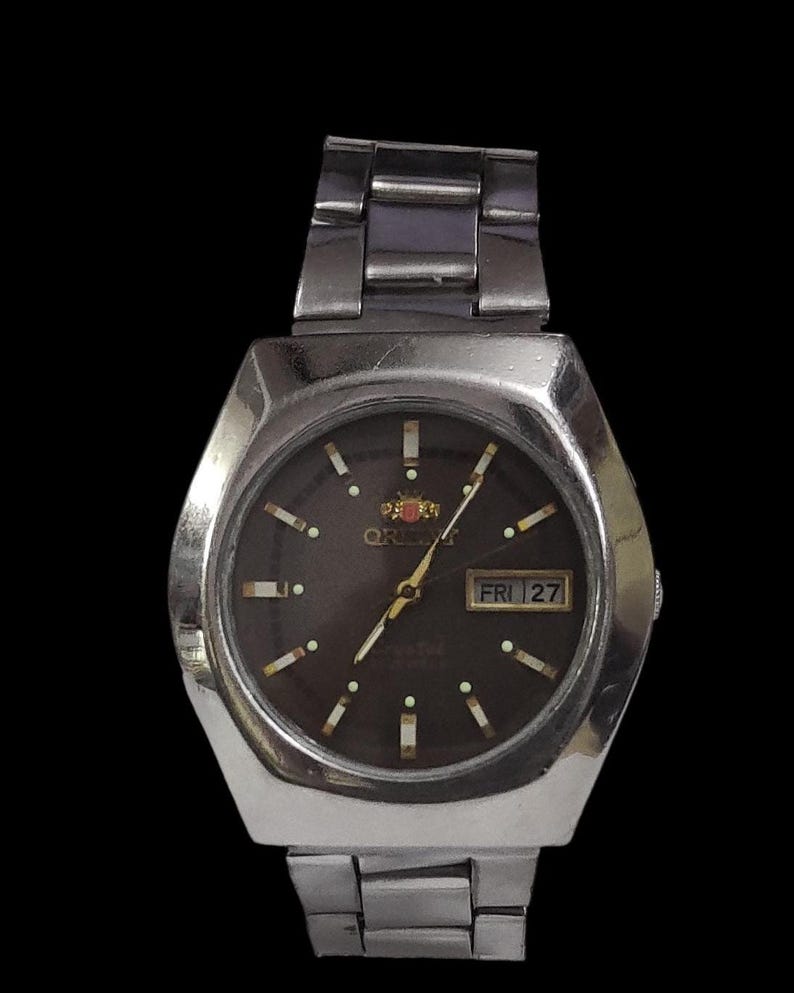 K&ouml;nnte beinhalten: Silberfarbene Orient-Uhr mit dunkelbraunem Zifferblatt. Die Uhr verf&uuml;gt &uuml;ber eine Tages- und Datumsanzeige, Leuchtmarkierungen und ein silbernes Metallarmband. Auf dem Zifferblatt steht "Orient" und "Crystal".