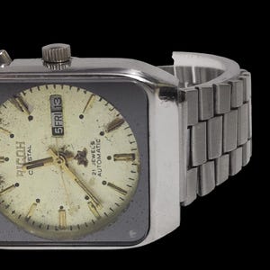 Pu&ograve; includere: Orologio automatico Ricoh Crystal vintage con cassa e cinturino color argento. Il quadrante &egrave; color crema con dettagli neri e oro, incluso il display del giorno e della data. L'orologio ha un quadrante quadrato e un cinturino in metallo.