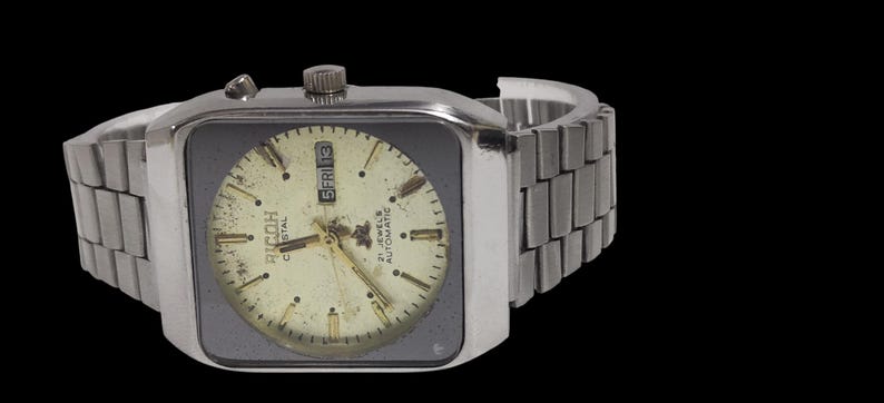 Pu&ograve; includere: Orologio automatico Ricoh Crystal vintage con cassa e cinturino color argento. Il quadrante &egrave; color crema con dettagli neri e oro, incluso il display del giorno e della data. L'orologio ha un quadrante quadrato e un cinturino in metallo.