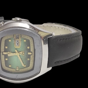 Puede incluir: Un reloj de pulsera vintage con caja plateada y correa de cuero negro. La esfera del reloj tiene un degradado verde y dorado, con agujas e indicadores dorados. El d&iacute;a se muestra como "LUN 15".