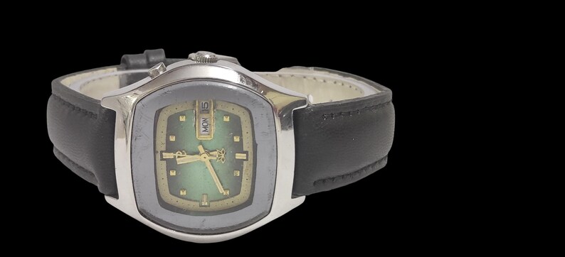 Puede incluir: Un reloj de pulsera vintage con caja plateada y correa de cuero negro. La esfera del reloj tiene un degradado verde y dorado, con agujas e indicadores dorados. El d&iacute;a se muestra como "LUN 15".