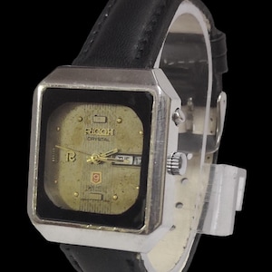 Może przedstawiać: Zegarek Ricoh Crystal w stylu vintage z kwadratową, srebrną kopertą i złotą tarczą. Zegarek posiada czarny sk&oacute;rzany pasek i wyświetla dzień i datę. Klasyczny zegarek.