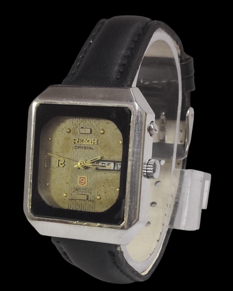 Może przedstawiać: Zegarek Ricoh Crystal w stylu vintage z kwadratową, srebrną kopertą i złotą tarczą. Zegarek posiada czarny sk&oacute;rzany pasek i wyświetla dzień i datę. Klasyczny zegarek.