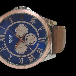 Może przedstawiać: Zegarek na rękę Casio z niebieską tarczą, srebrną kopertą i brązowym sk&oacute;rzanym paskiem. Zegarek posiada rzymskie cyfry i trzy sub-tarcze. Ramka jest srebrna z pierścieniem w kolorze r&oacute;żowego złota. Pasek ma szerokość około 2 cm.