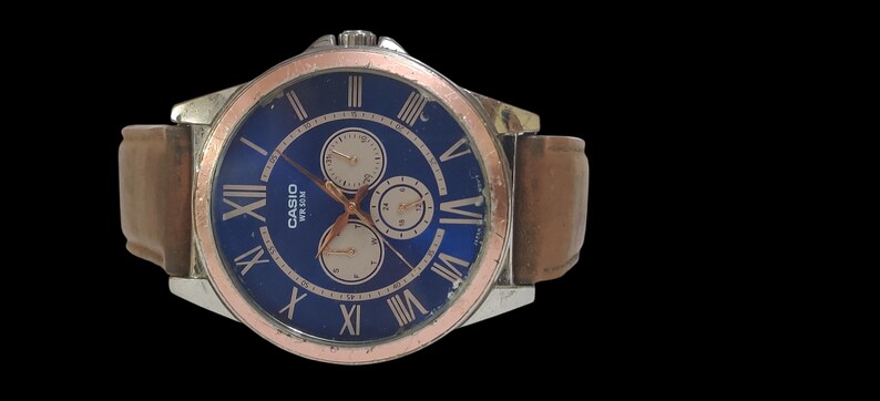 Może przedstawiać: Zegarek na rękę Casio z niebieską tarczą, srebrną kopertą i brązowym sk&oacute;rzanym paskiem. Zegarek posiada rzymskie cyfry i trzy sub-tarcze. Ramka jest srebrna z pierścieniem w kolorze r&oacute;żowego złota. Pasek ma szerokość około 2 cm.