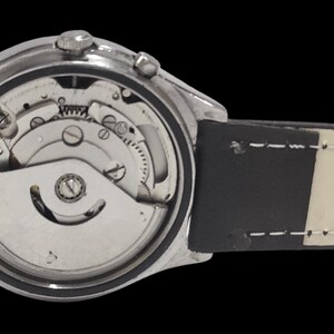 Puede incluir: Primer plano de un reloj de pulsera plateado con movimiento mec&aacute;nico visible. El reloj tiene una correa de cuero en blanco y negro. Se exponen los intrincados mecanismos internos del reloj, mostrando engranajes y otros componentes.