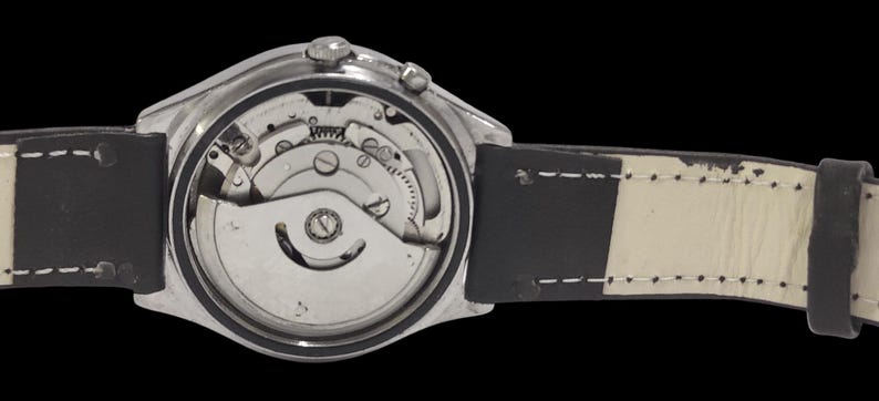 Puede incluir: Primer plano de un reloj de pulsera plateado con movimiento mec&aacute;nico visible. El reloj tiene una correa de cuero en blanco y negro. Se exponen los intrincados mecanismos internos del reloj, mostrando engranajes y otros componentes.
