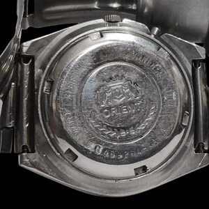 K&ouml;nnte beinhalten: Nahaufnahme einer silberfarbenen Orient-Uhr. Die R&uuml;ckseite der Uhr zeigt das Orient-Logo, Text und eine Seriennummer. Das Uhrenarmband ist ebenfalls silberfarben. Die Uhr ist vor einem schwarzen Hintergrund platziert.