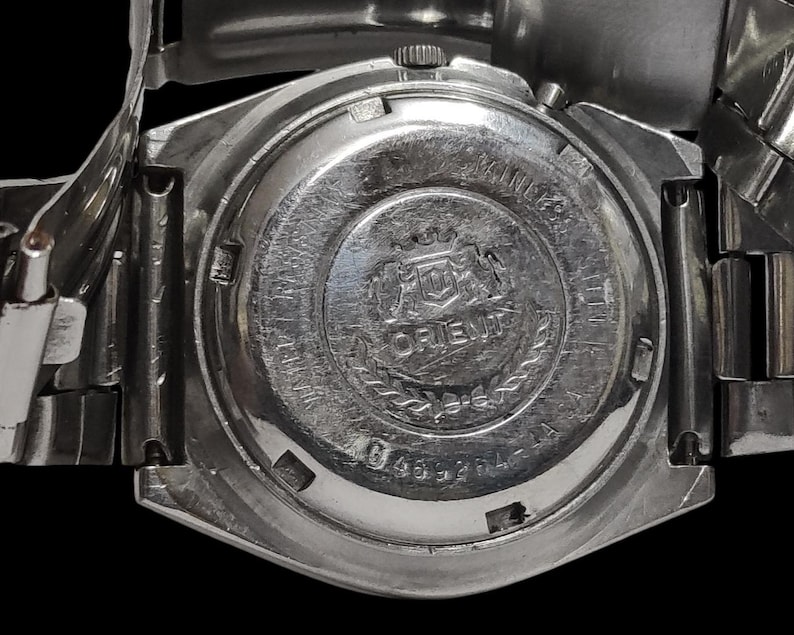K&ouml;nnte beinhalten: Nahaufnahme einer silberfarbenen Orient-Uhr. Die R&uuml;ckseite der Uhr zeigt das Orient-Logo, Text und eine Seriennummer. Das Uhrenarmband ist ebenfalls silberfarben. Die Uhr ist vor einem schwarzen Hintergrund platziert.