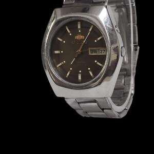 K&ouml;nnte beinhalten: Eine silberfarbene Orient-Uhr mit dunkelbraunem Zifferblatt und Metallarmband. Das Zifferblatt zeigt den Tag und das Datum an. Die Uhr hat ein rechteckiges Geh&auml;use und ein silberfarbenes Metallarmband.