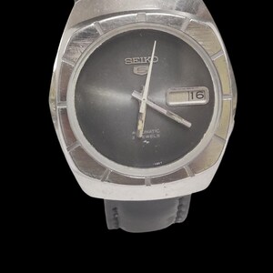 May include: A vintage Seiko watch with a black dial and silver-tone case. The watch features a date display at the 3 o'clock position and a black leather strap. The watch face has the word "SEIKO" and "AUTOMATIC 2 JEWELS" printed on it.