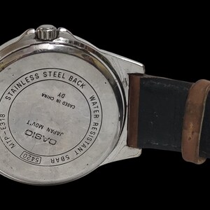 Może przedstawiać: Srebrny zegarek Casio z brązowym sk&oacute;rzanym paskiem. Widoczny jest tył zegarka z napisami, w tym "Stainless Steel Back" i "Water Resistant 5BAR". Pasek jest czarny wewnątrz i brązowy na zewnątrz.