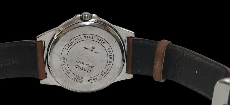 Może przedstawiać: Srebrny zegarek Casio z brązowym sk&oacute;rzanym paskiem. Widoczny jest tył zegarka z napisami, w tym "Stainless Steel Back" i "Water Resistant 5BAR". Pasek jest czarny wewnątrz i brązowy na zewnątrz.