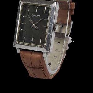 Puede incluir: Un reloj Sonata rectangular con esfera oscura y caja plateada. El reloj tiene una correa de cuero marr&oacute;n con un patr&oacute;n texturizado. El reloj est&aacute; sobre un fondo negro.