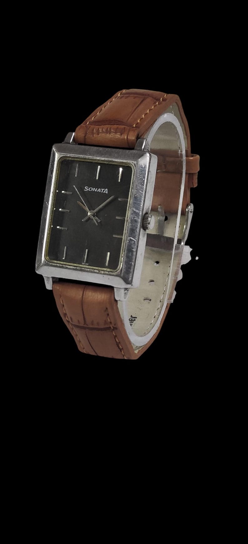 Puede incluir: Un reloj Sonata rectangular con esfera oscura y caja plateada. El reloj tiene una correa de cuero marr&oacute;n con un patr&oacute;n texturizado. El reloj est&aacute; sobre un fondo negro.