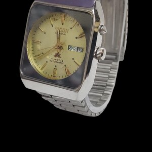 Pu&ograve; includere: Un orologio da polso automatico Ricoh Crystal vintage con cassa rettangolare color argento e cinturino a maglie argentate. Il quadrante dell'orologio &egrave; dorato con lancette e indici neri e un display giorno/data.