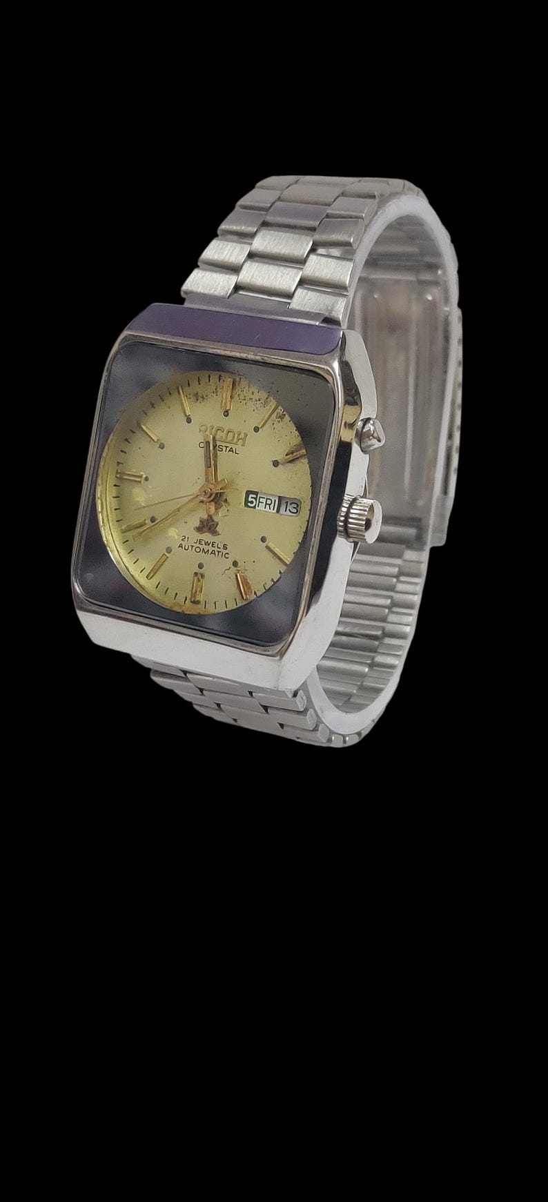 Pu&ograve; includere: Un orologio da polso automatico Ricoh Crystal vintage con cassa rettangolare color argento e cinturino a maglie argentate. Il quadrante dell'orologio &egrave; dorato con lancette e indici neri e un display giorno/data.