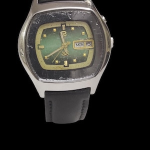 Puede incluir: Un reloj de pulsera vintage con caja plateada y correa de cuero negro. La esfera del reloj es verde con manecillas e indicadores dorados. Se muestra el d&iacute;a y la fecha. El reloj est&aacute; sobre un fondo negro.