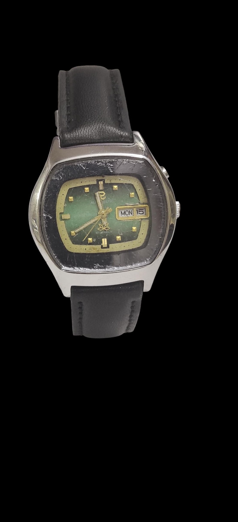 Puede incluir: Un reloj de pulsera vintage con caja plateada y correa de cuero negro. La esfera del reloj es verde con manecillas e indicadores dorados. Se muestra el d&iacute;a y la fecha. El reloj est&aacute; sobre un fondo negro.