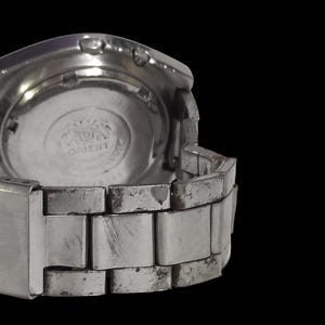 K&ouml;nnte beinhalten: Eine silberfarbene Armbanduhr mit Metallarmband, von hinten betrachtet. Das Zifferblatt ist sichtbar, mit dem Markennamen "Orient" aufgedruckt. Das Armband weist Gebrauchsspuren auf.