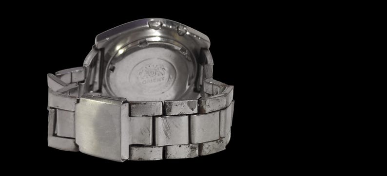 K&ouml;nnte beinhalten: Eine silberfarbene Armbanduhr mit Metallarmband, von hinten betrachtet. Das Zifferblatt ist sichtbar, mit dem Markennamen "Orient" aufgedruckt. Das Armband weist Gebrauchsspuren auf.