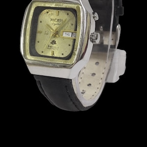 Pode incluir: Relógio de pulso vintage com caixa prateada e mostrador dourado. O relógio possui uma pulseira de couro preta e exibe o dia e a data. O mostrador do relógio tem a marca "Michel" e a palavra "Crystal".