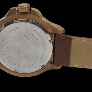 Peut inclure: Gros plan d'une montre dor&eacute;e avec un bracelet en cuir marron. Le cadran de la montre est argent&eacute; avec du texte noir. La montre a une couronne textur&eacute;e et un bracelet en cuir marron.