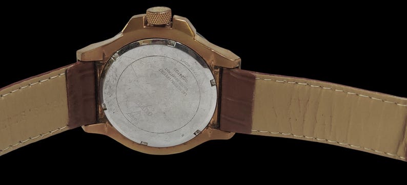 Peut inclure: Gros plan d'une montre dor&eacute;e avec un bracelet en cuir marron. Le cadran de la montre est argent&eacute; avec du texte noir. La montre a une couronne textur&eacute;e et un bracelet en cuir marron.
