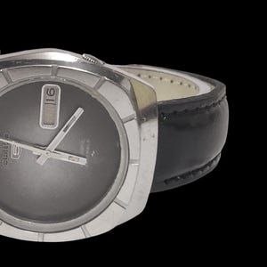 May include: A silver-toned wristwatch with a dark gray face and a black leather band. The watch has a rectangular face with silver hands and a date display at the 3 o'clock position. The band is stitched and has a buckle closure.