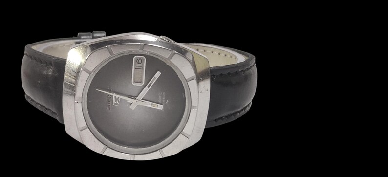 May include: A silver-toned wristwatch with a dark gray face and a black leather band. The watch has a rectangular face with silver hands and a date display at the 3 o'clock position. The band is stitched and has a buckle closure.