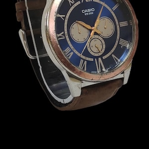 Może przedstawiać: Zegarek na rękę Casio z niebieską tarczą, srebrną kopertą i brązowym sk&oacute;rzanym paskiem. Tarcza zegarka posiada rzymskie cyfry i trzy dodatkowe tarcze. Zegarek jest prezentowany na czarnym tle.