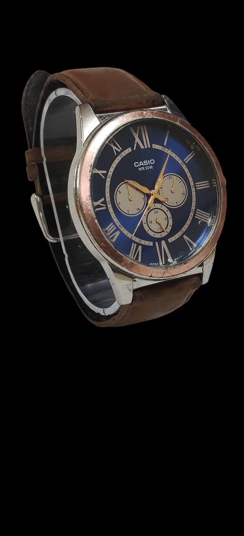 Może przedstawiać: Zegarek na rękę Casio z niebieską tarczą, srebrną kopertą i brązowym sk&oacute;rzanym paskiem. Tarcza zegarka posiada rzymskie cyfry i trzy dodatkowe tarcze. Zegarek jest prezentowany na czarnym tle.