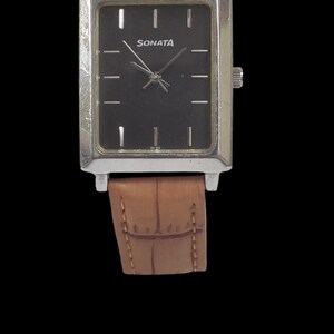 Puede incluir: Un reloj Sonata rectangular con esfera negra y caja plateada. El reloj tiene una correa de cuero marr&oacute;n. La esfera del reloj tiene marcadores de hora plateados y el nombre de la marca "Sonata" en blanco.