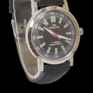 Może przedstawiać: Zegarek na rękę Fortis w stylu vintage z czarną tarczą, srebrną kopertą i czarnym skórzanym paskiem. Tarcza zegarka wyświetla nazwę marki "FORTIS" oraz tekst "17 JEWELS SHOCK PROOF". Zegarek ma zielone akcenty i sekundnik.