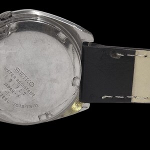 May include: A stainless steel Seiko watch with a black leather strap. The watch face is silver with the text "SEIKO WATER RESISTANT 5 9 7 2 7 JAPAN-A 7019-7070". The strap is black with white stitching.