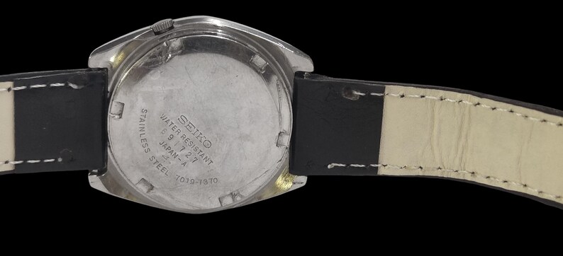 May include: A stainless steel Seiko watch with a black leather strap. The watch face is silver with the text "SEIKO WATER RESISTANT 5 9 7 2 7 JAPAN-A 7019-7070". The strap is black with white stitching.