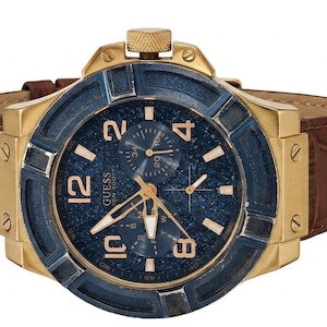 Peut inclure: Une montre Guess dor&eacute;e avec un cadran bleu et un bracelet en cuir marron. Le cadran de la montre pr&eacute;sente des aiguilles et des chiffres dor&eacute;s. La montre a une lunette bleu fonc&eacute; textur&eacute;e et une couronne dor&eacute;e.