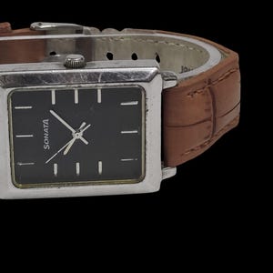 Puede incluir: Un reloj Sonata rectangular con esfera negra y caja plateada. El reloj tiene una correa de cuero marr&oacute;n. La esfera muestra el nombre de la marca y marcadores de hora sencillos. El reloj est&aacute; sobre un fondo negro.
