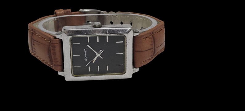 Puede incluir: Un reloj Sonata rectangular con esfera negra y caja plateada. El reloj tiene una correa de cuero marr&oacute;n. La esfera muestra el nombre de la marca y marcadores de hora sencillos. El reloj est&aacute; sobre un fondo negro.