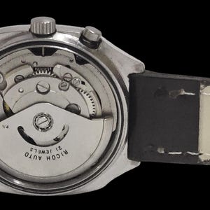 May include: A close-up view of a silver wristwatch with a black leather strap. The watch's back is open, revealing the internal mechanical workings, including gears and a rotor. The watch face reads "RICOH AUTO 21 JEWELS".