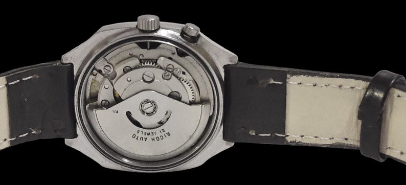 May include: A close-up view of a silver wristwatch with a black leather strap. The watch's back is open, revealing the internal mechanical workings, including gears and a rotor. The watch face reads "RICOH AUTO 21 JEWELS".