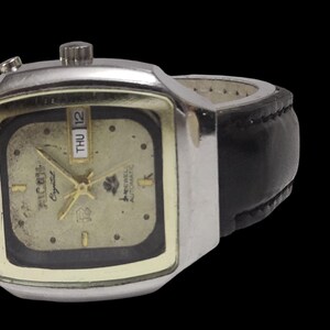 May include: A vintage wristwatch with a silver square case and a gold face. It has a black leather strap and shows the day and date. The watch face has the text "RICOH Crystal" and a logo.