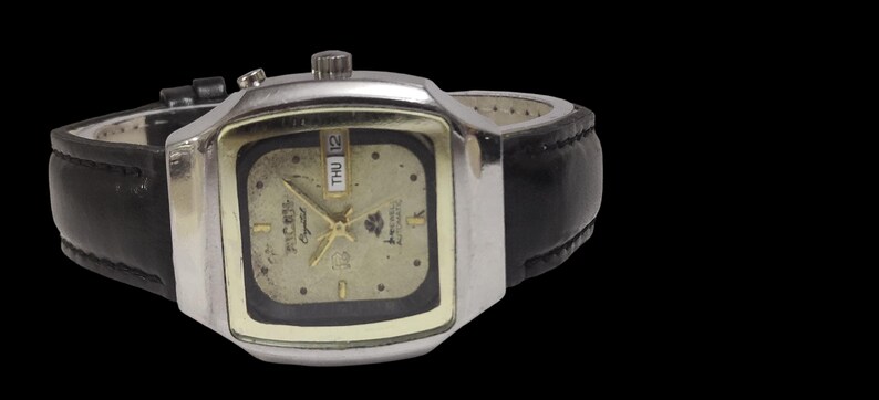 May include: A vintage wristwatch with a silver square case and a gold face. It has a black leather strap and shows the day and date. The watch face has the text "RICOH Crystal" and a logo.