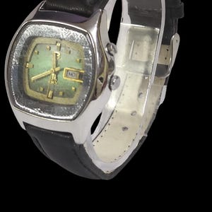 Puede incluir: Un reloj de pulsera vintage con una caja rectangular plateada y una esfera verde. El reloj presenta agujas e indicadores dorados, y una pantalla de d&iacute;a y fecha. Tiene una correa de cuero negro.